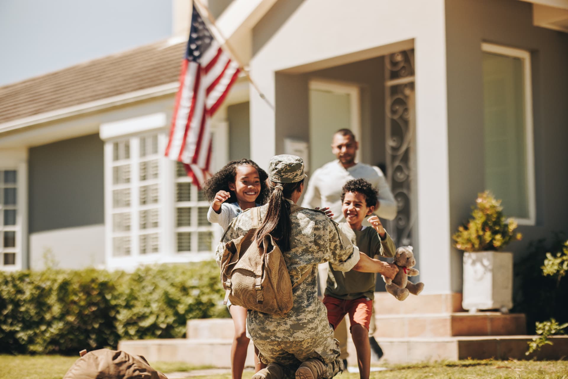 Service member in uniform kneeling to hug her children on the front lawn after returning home