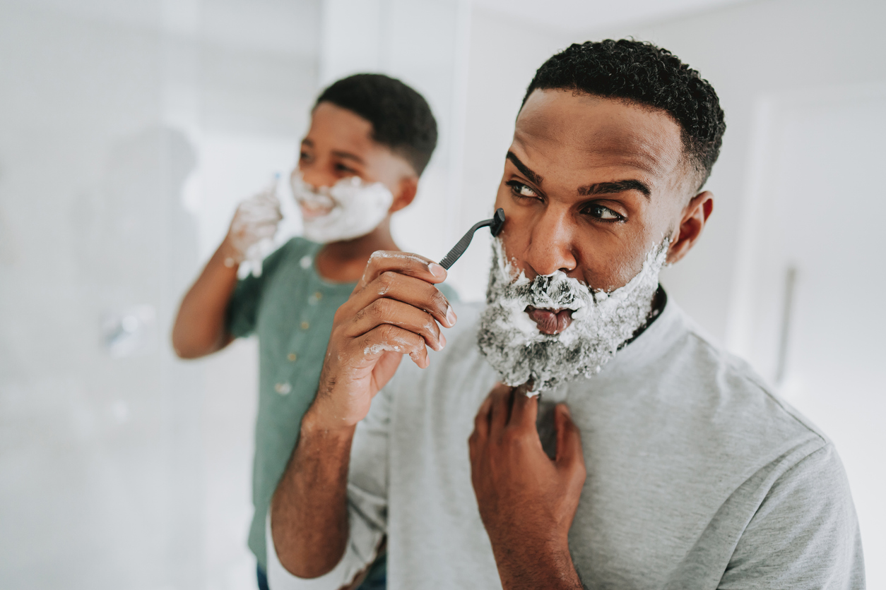 Man shaving at a bathroom mirror while his young son shaves alongside him