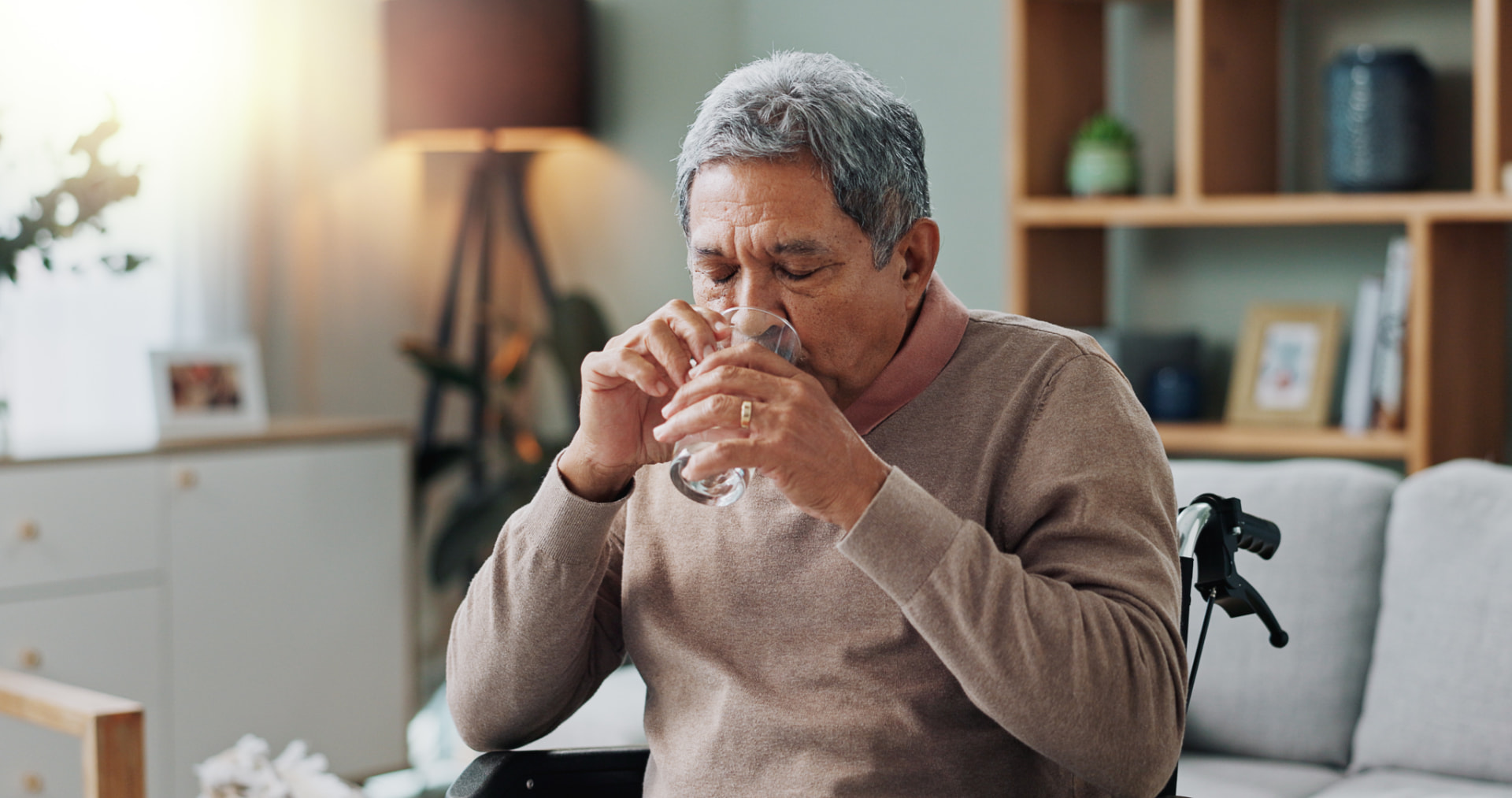 Older man seated indoors, holding a glass of water with both hands, with a walking cane beside him