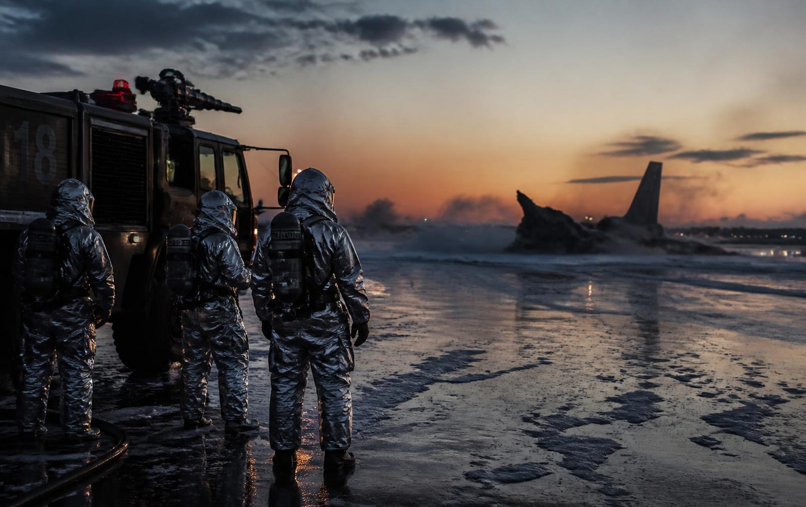 Military aircraft rescue firefighting crew in silver proximity suits beside a crash truck on a wet airfield at dusk